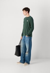 Groene lange mouwen shirt met een klein logo, versleten lichtblauwe wijde jeans, beige sneakers en een zwarte tote bag.