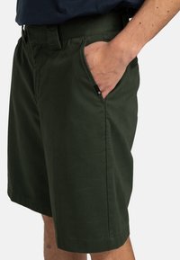 Grüne Baumwollshorts mit lockerer Passform, ausgestattet mit seitlichen Taschen und einem Etikett. Der Stoff hat eine glatte, atmungsaktive Textur.