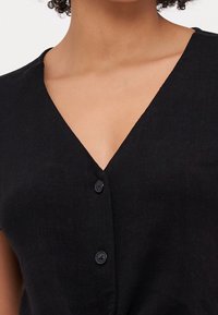 Donna che indossa un top nero senza maniche a V con due bottoni sul davanti, che mette in mostra la zona delle clavicole e del collo.