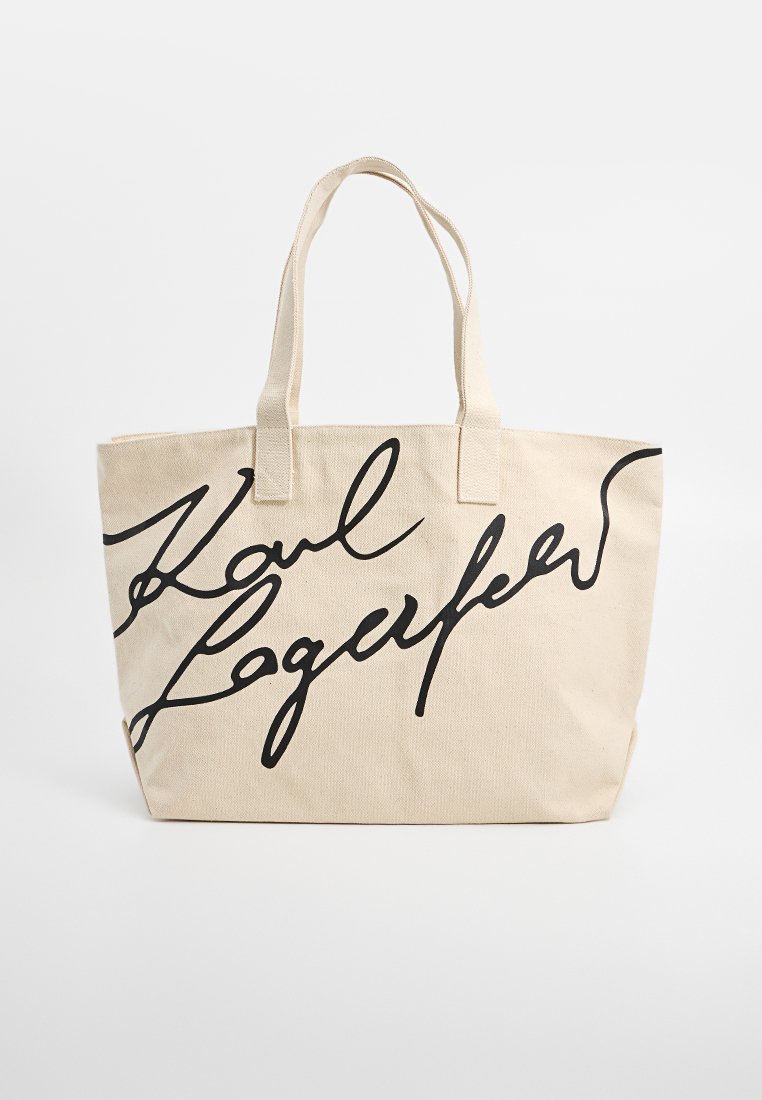 Sac en toile beige avec une écriture cursive noire inscrivant "Karl Lagerfeld" et deux poignées sur fond blanc.