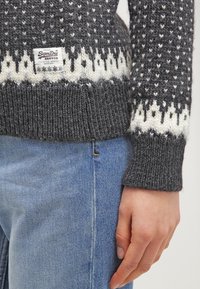 Harmaa neulepaita, jossa on valkoisia geometrisia kuvioita, resorit hihansuissa ja helmassa, edessä näkyvä merkintälappu. Päällä vaaleansiniset farkut.