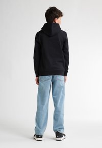 Felpa nera con cappuccio ampio, abbinata a jeans chiari a gamba larga e sneakers nere con dettagli bianchi e logo.