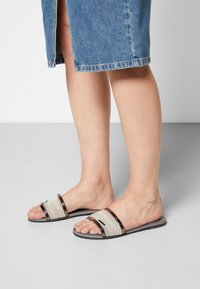 Piedi che indossano sandali a scivolo con bordi argentati e cinturini in tessuto bianco con motivi, abbinati a una gonna di jeans blu con uno spacco sul davanti.