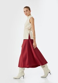 Beige mouwloze top met zijstrikjes, gecombineerd met een vloeiende rode satijnen rok en witte kniehoge spitse laarzen. Eenvoudige, elegante silhouet.