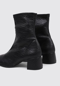 Botas de cuero negras hasta el tobillo con un acabado texturizado, punta redondeada y tacón bajo de media altura. Presentan un diseño alto en el tobillo y un detalle minimalista.