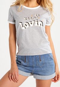 Šedé tričko s krátkým rukávem a bílými lemy, na kterém je nápis "KEEP Lovin’" napsaný v různých typech písma. K tričku jsou dolní části oblečení - džínové šortky, což ukazuje na ležérní styl.