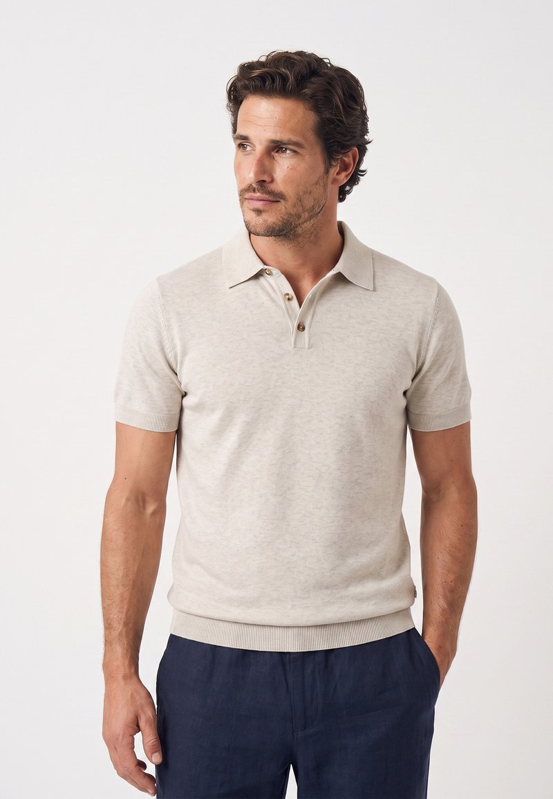Man met donker haar, gekleed in een beige polo met korte mouwen en donkerblauwe broek, staat met één hand in de zak, kijkt naar links.