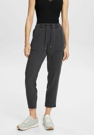 Vrouw draagt zwart mouwloos topje, antracietgrijze broek met trekkoord en grote voorzakken en lichtgrijze sneakers, staand op een witte achtergrond.