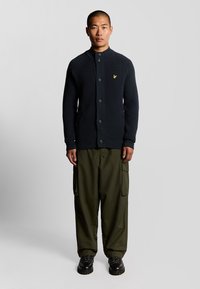 Cardigan lavorato a maglia blu navy con colletto alto e chiusura a bottoni, abbinato a pantaloni cargo verde oliva. Calzature nere robuste completano il look.