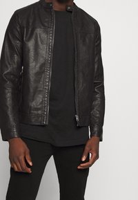Veste en cuir noir avec un col montant, fermeture éclair et poches latérales ; portée par-dessus une chemise noire, texture lisse, ajustement sur mesure.