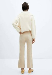 Crème-kleurige oversized trui met een hoge kraag, gecombineerd met beige flare broek en lichtbruine schoenen. Gladde textuur, minimale details.