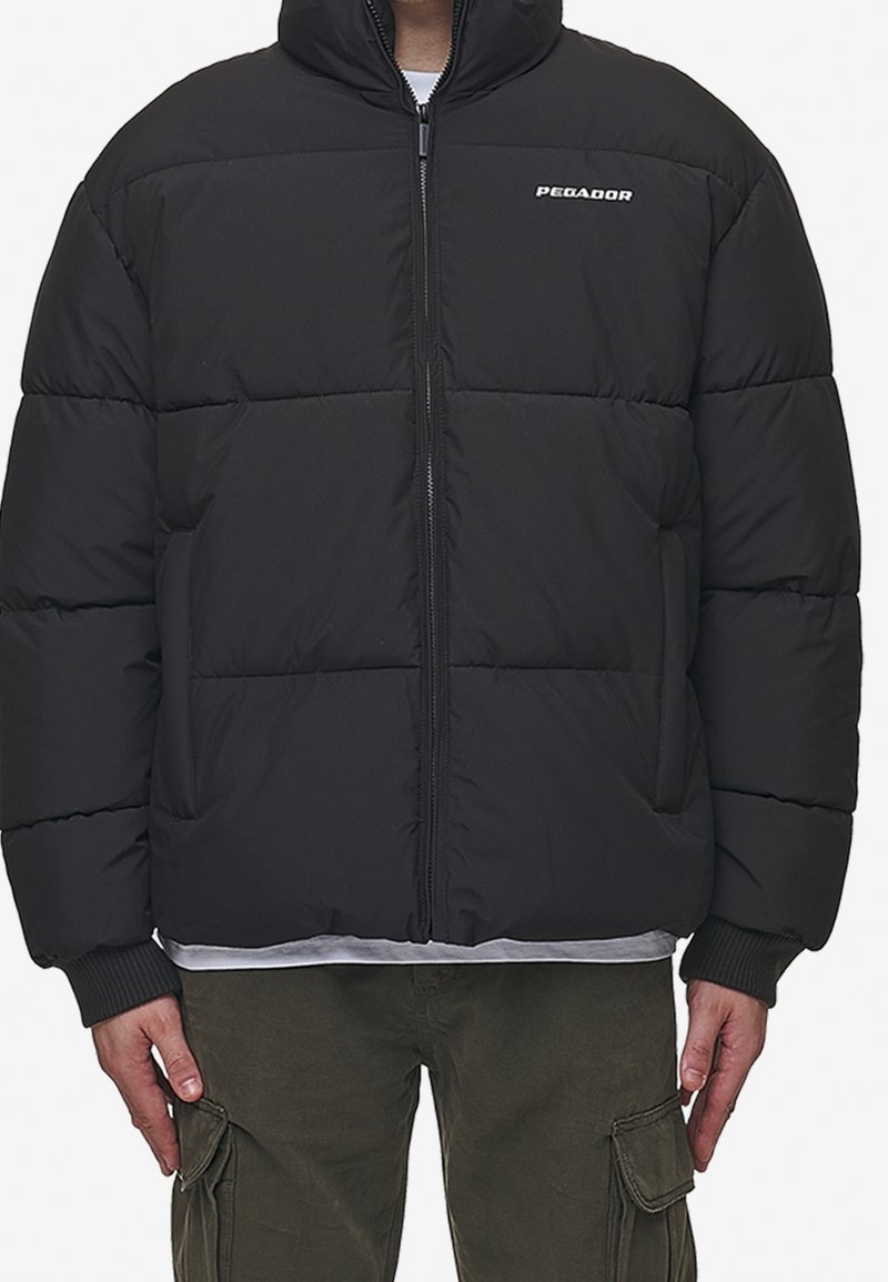 Sort polstret jakke med høj krave, forreste lynlås, elastiske manchetter og et PEGAADOR-logo på brystet. Har en quiltet struktur.