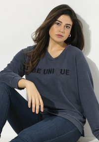Dunkelgraues, oversized Sweatshirt mit schwarzem "BE UNIQUE"-Text, V-Ausschnitt, rohem Saum und langen Ärmeln, kombiniert mit dunklen Jeans.