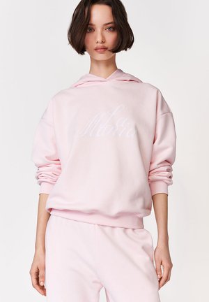 Młoda kobieta o krótkich ciemnych włosach, ubrana w jasnoróżową bluzę z kapturem i pasujące spodnie dresowe, stoi na jednolitym białym tle.