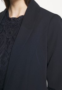 Námornický blazer se strukturálními rameny a hladkým povrchem, vrstvený přes tmavý krajkový top s výjimečnými vzory.