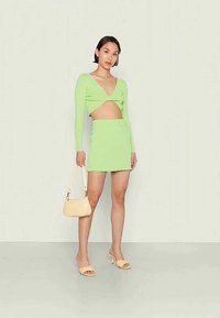 Top cropped verde de malha com mangas longas e um detalhe recortado, combinado com uma saia mini justa. Os acessórios incluem uma pequena mala amarela e sapatos de salto.