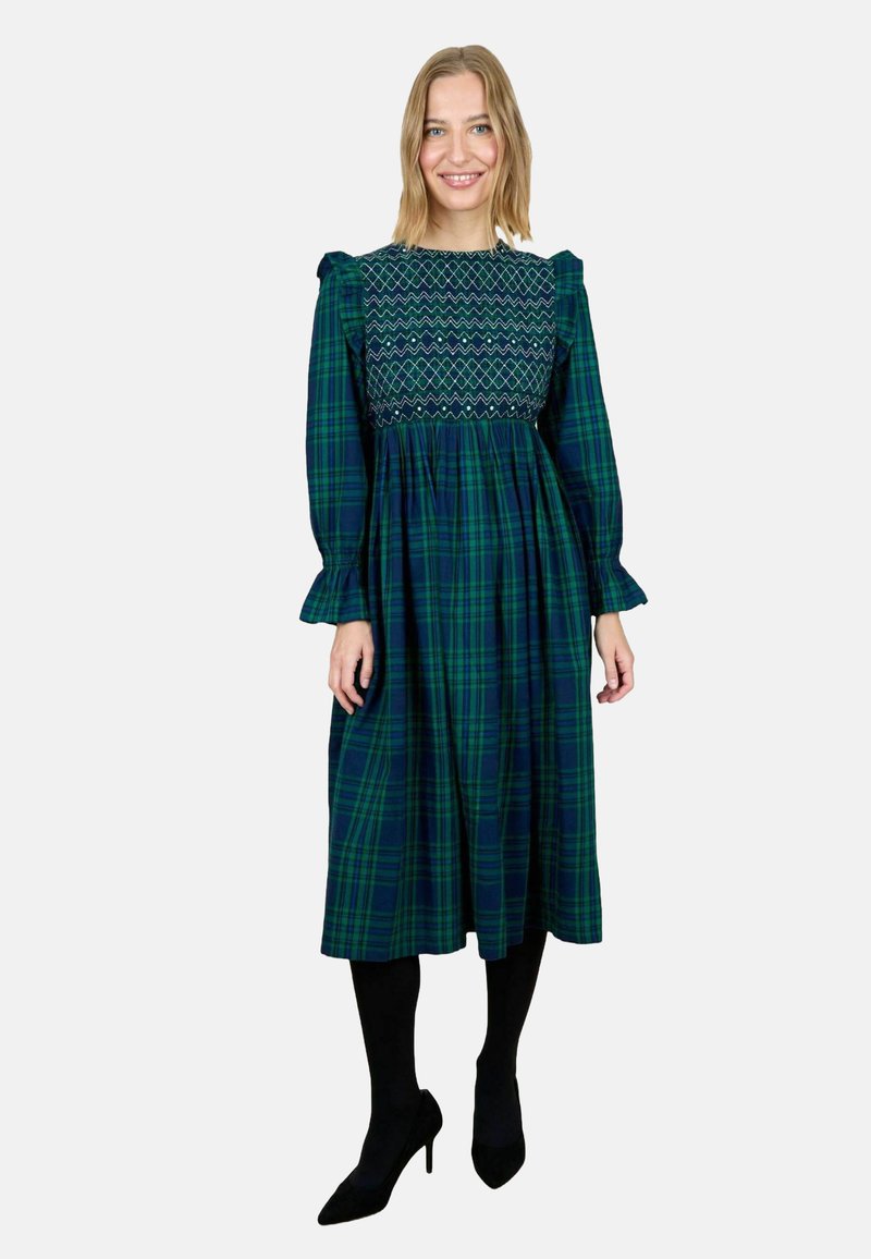 Femeie îmbrăcată într-o rochie midi în carouri verzi și bleumarin, cu mâneci lungi și puffate, un corp modelat cu elastic, colanți negri și pantofi cu vârf ascuțit negri, zâmbind.