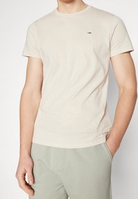Beige bavlněné tričko s kulatým výstřihem, krátkými rukávy a malou vyšitou nášivkou na hrudi. Hladká textura, fitted design.