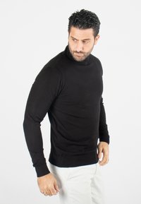 Pull en laine noir à manches longues avec col roulé en tissu tricoté lisse, design ajusté avec des poignets et un ourlet côtelés, associé à un pantalon de couleur claire.