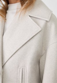 Light grey, textured wool-blend coat with a wide lapel, vertical front pockets, and structured shoulders. Features a soft, warm finish.