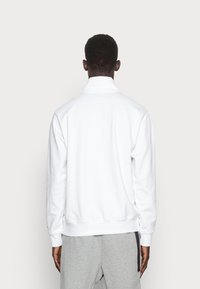 Sudadera blanca de manga larga y cuello alto, con puños y dobladillo de canalé. Textura suave y diseño minimalista, llevada sobre pantalones grises.