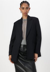 Blazer nero a righe con rever a punte e chiusura a un bottone, indossato sopra un top grigio trasparente e abbinato a una gonna di pelle nera.