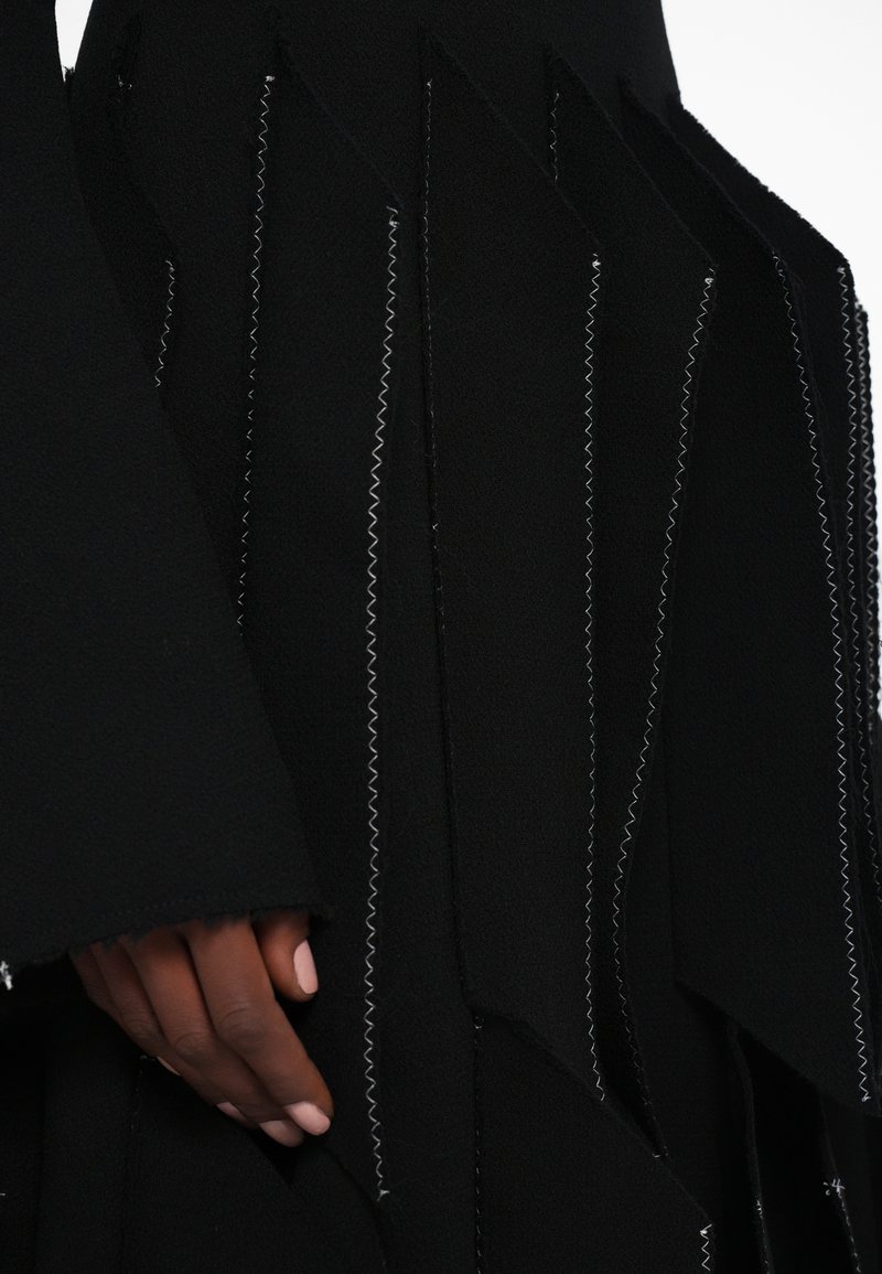 Black garment featuring an asymmetric design with textured fabric, zigzag stitching, and layered sections. Close-up of hand gripping the fabric.