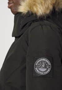 Svart jacka med huva i konstpäls. Har en rund, broderad logotypmärke på vänster axel som visar berg och vatten.