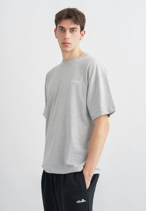 Młody mężczyzna o krótkich brązowych włosach, ubrany w szarą koszulkę ellesse oraz czarne spodnie ellesse, stoi z jedną ręką w kieszeni.