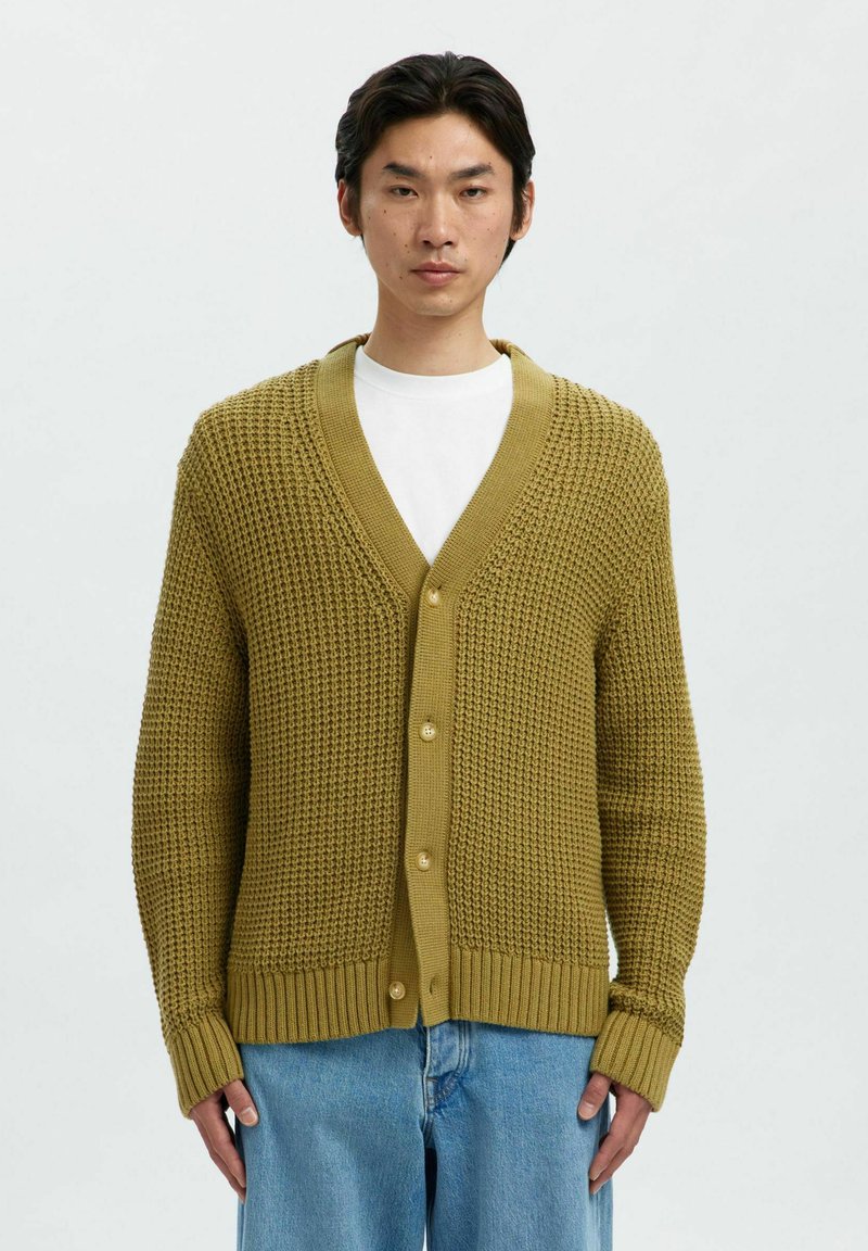 Man i en senapsgul, strukturerad stickad cardigan över en vit skjorta med ljusblå jeans, stående mot en enfärgad ljus bakgrund.