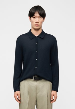 Mand iført en langærmet navy skjorte med knapper, stoppet ned i beige plisserede bukser med sort bælte, stående mod en ensfarvet baggrund.