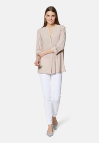 Helle beige Bluse mit V-Ausschnitt und knöpfbaren Ärmeln, kombiniert mit weißen Slim-Fit-Hosen und silbernen Ballerinas. Einfaches und elegantes Design.