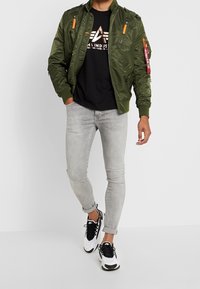 Olivově zelená bomber bunda s oranžovými akcenty, černé tričko s grafickým potiskem, světle šedé slim-fit džíny a černo-bílé sportovní tenisky.
