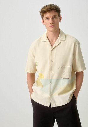 Ung mand med lysebrunt hår bærer en cremefarvet kortærmet skjorte med et motiv af en sejlbåd, sol, fugle og strand, kombineret med sorte bukser.