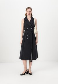 Μαύρο αμάνικο παλτό φόρεμα με ζώνη στη μέση, διπλά κουμπιά και μήκος μέχρι το γόνατο σε Α γραμμή. Φορεμένο με μαύρες γόβες και λευκή τσάντα.