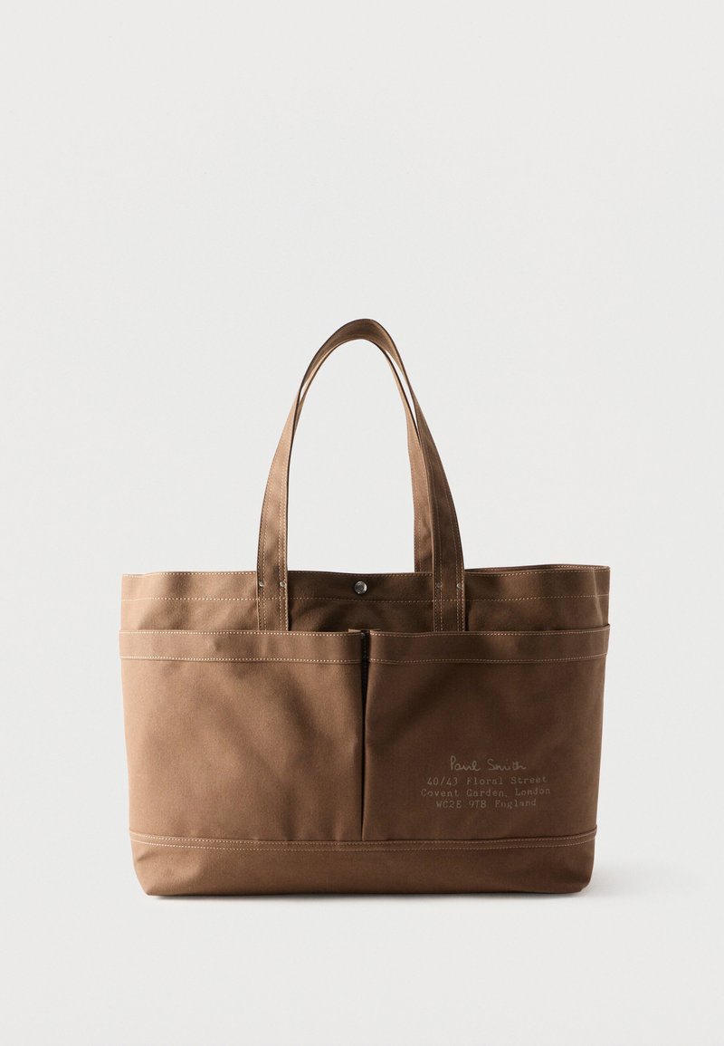 Sac fourre-tout en toile marron avec doubles poignées, poches avant et texte imprimé discret sur la poche inférieure droite.