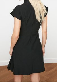 Une personne aux longs cheveux blonds porte une robe noire à manches courtes, vue de dos contre un mur clair et un sol en bois.