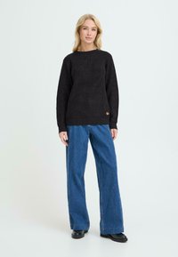 Suéter negro de punto con un patrón texturizado y puños acanalados, combinado con jeans de denim azul de pierna ancha y botas negras hasta el tobillo.