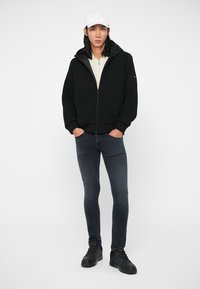 Black puffer jacket with ribbed texture, hood, and zip closure. Worn over a beige top and dark skinny jeans. Black sneakers complete the look.