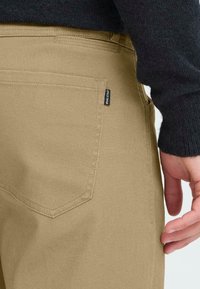 Beige bomuldsbukser med en struktureret stof, der har en baglomme med et sort mærke og en skræddersyet kant, delvist vist sammen med en mørk sweater.