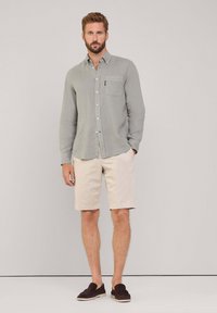 Camicia a maniche lunghe grigia chiara con bottoni e taschino sul petto, abbinata a pantaloni corti beige e scarpe scivolate scure, presentata su uno sfondo neutro.