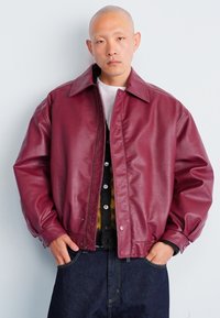 Bordeaux leren jas met een ontspannen pasvorm, grote kraag en drukknoopsluiting, gedragen over een zwart en geel gestreept trui, gecombineerd met donkere denim.