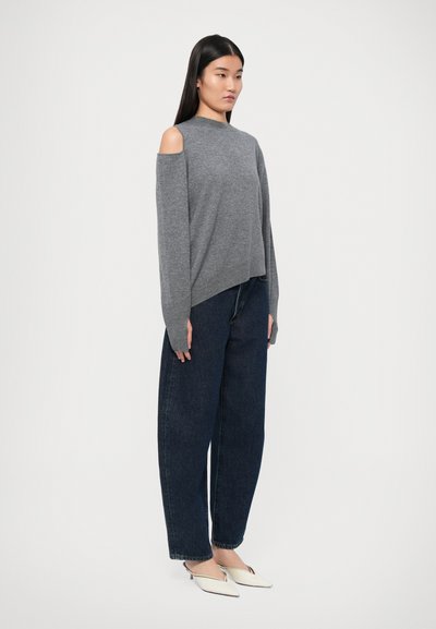 Maglione grigio a maglia con design a spalle scoperte, abbinato a jeans in denim blu scuro a vestibilità ampia e tacchi bianchi a punta.