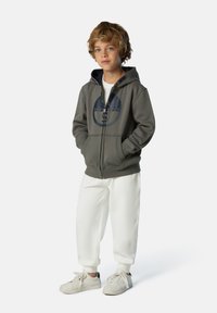 Grauer Reißverschluss-Hoodie mit einem runden Logo, kombiniert mit weißen Jogginghosen und hellen Sneakern, die ein bequemes, lässiges Design aufweisen.