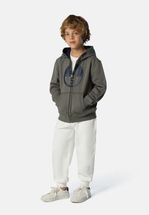 Felpa grigia con zip e logo circolare, abbinata a pantaloni della tuta bianchi e sneakers chiare, caratterizzata da un design comodo e casual.