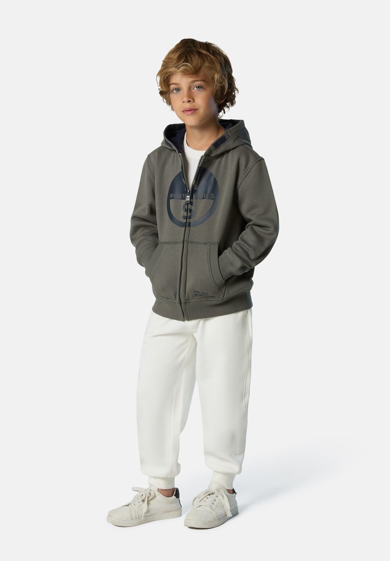 Grauer Reißverschluss-Hoodie mit einem runden Logo, kombiniert mit weißen Jogginghosen und hellen Sneakern, die ein bequemes, lässiges Design aufweisen.