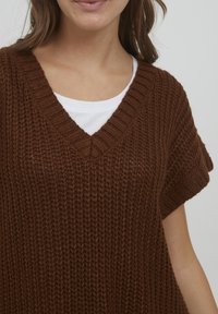 Top en tricot marron avec un décolleté en V et des manches courtes, présentant une coupe ample et un motif texturé. Porté sur une chemise blanche.