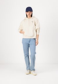 Felpa crema con tasca frontale, jeans azzurri chiari e berretto da baseball blu navy. Tessuto testurizzato, vestibilità casual e palette di colori neutri.