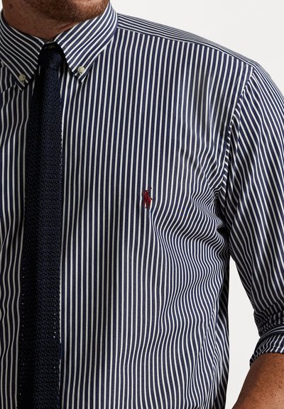 Μπλε και λευκή ριγέ πουκάμισο με κουμπιά, που διαθέτει ένα μικρό κόκκινο λογότυπο, συνδυασμένο με μια υφασμάτινη μπλε γραβάτα. Κατασκευασμένο από βαμβακερό ύφασμα με απαλή υφή.