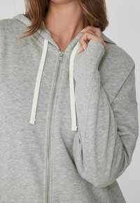 Grau zip-up Hoodie aus weichem Material, mit einer großen Kapuze, hellen Kordeln und einem lässigen Schnitt mit angenehmer Textur.